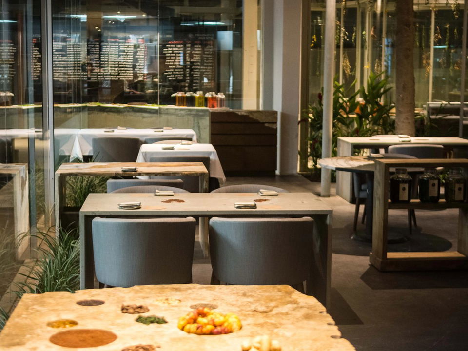 Interior of a modern, upscale restaurant with concrete tables, grey chairs, glass walls, and lush plants, featuring a menu board reflection.