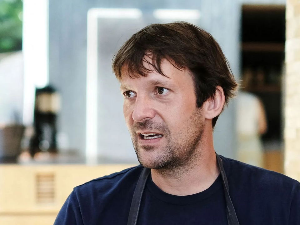 Chef speaking in a modern kitchen setting with blurred background activity.