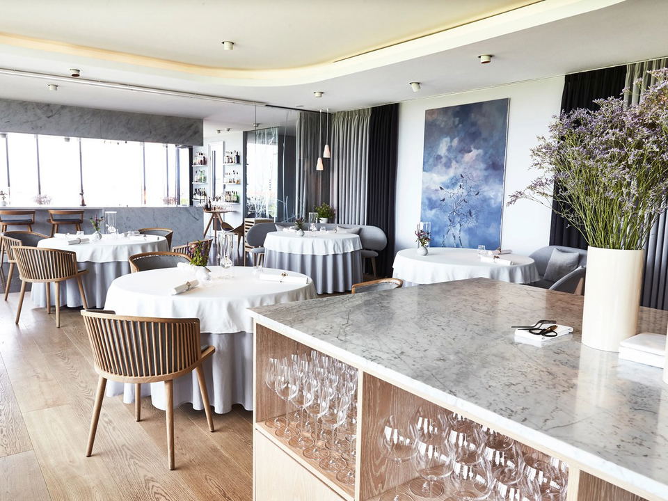 An elegant fine-dining restaurant interior with round tables draped in white and grey linen, wooden spindle-back chairs, a marble service counter, a