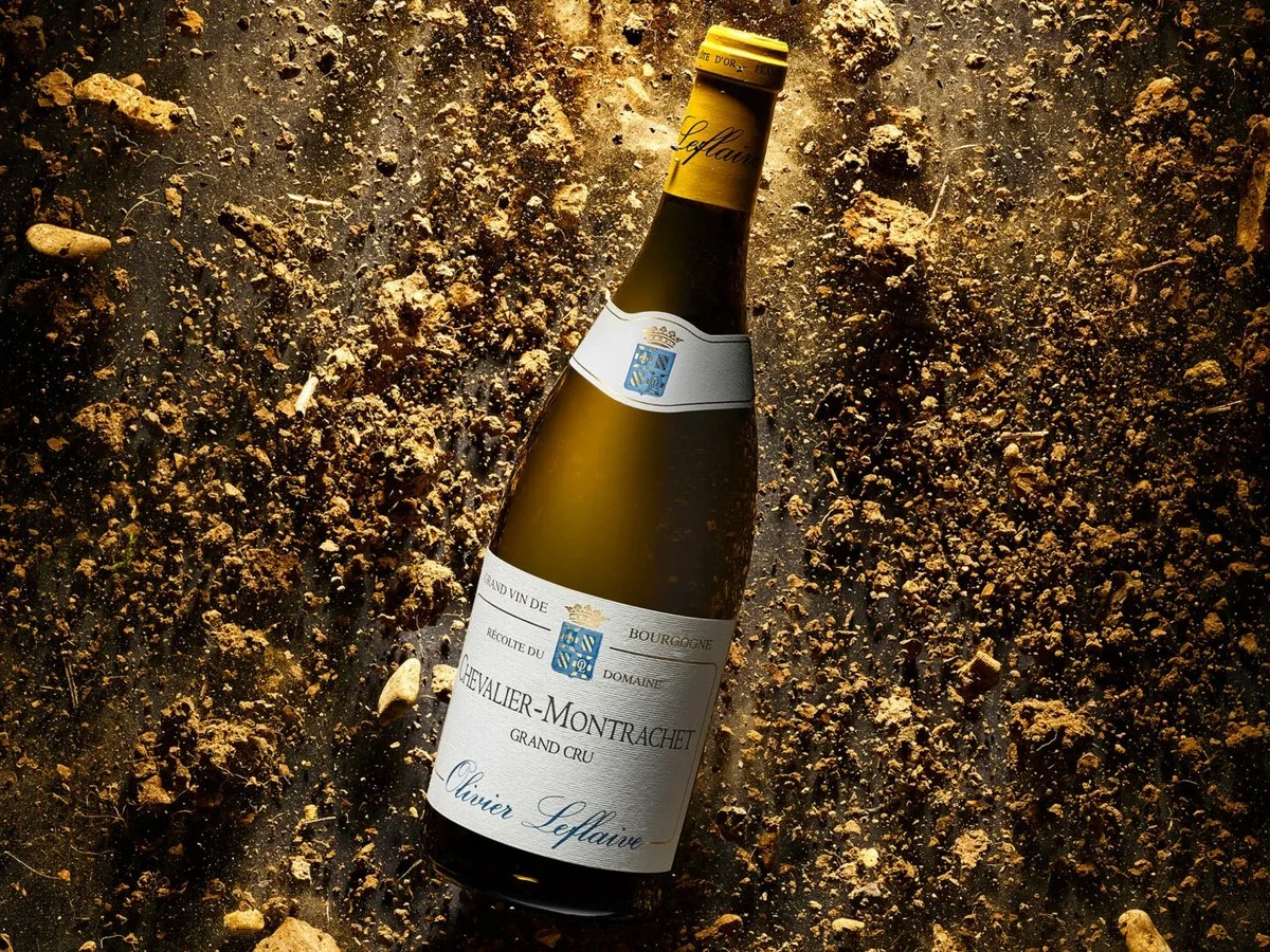A bottle of Olivier Leflaive Chevalier-Montrachet Grand Cru white wine, with a yellow foil top, lying on a dark, textured surface covered in soil and