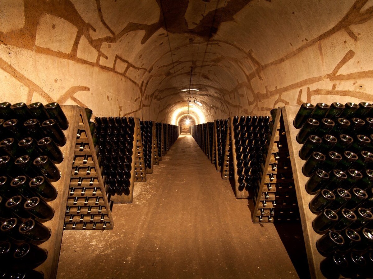 A long, dimly lit underground tunnel cellar with rows of champagne bottles stored on wooden riddling racks, angled neck-down.