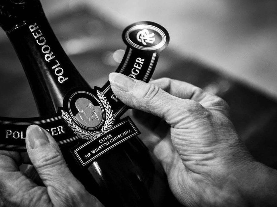 A pair of hands holds a dark bottle of Pol Roger Cuvée Sir Winston Churchill champagne, with the label clearly visible.
