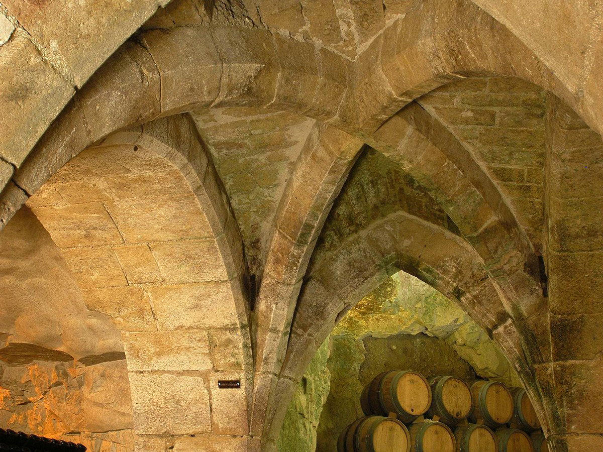 A medieval stone-vaulted underground cellar with Gothic ribbed arches, stacked wine bottles, and oak barrels.