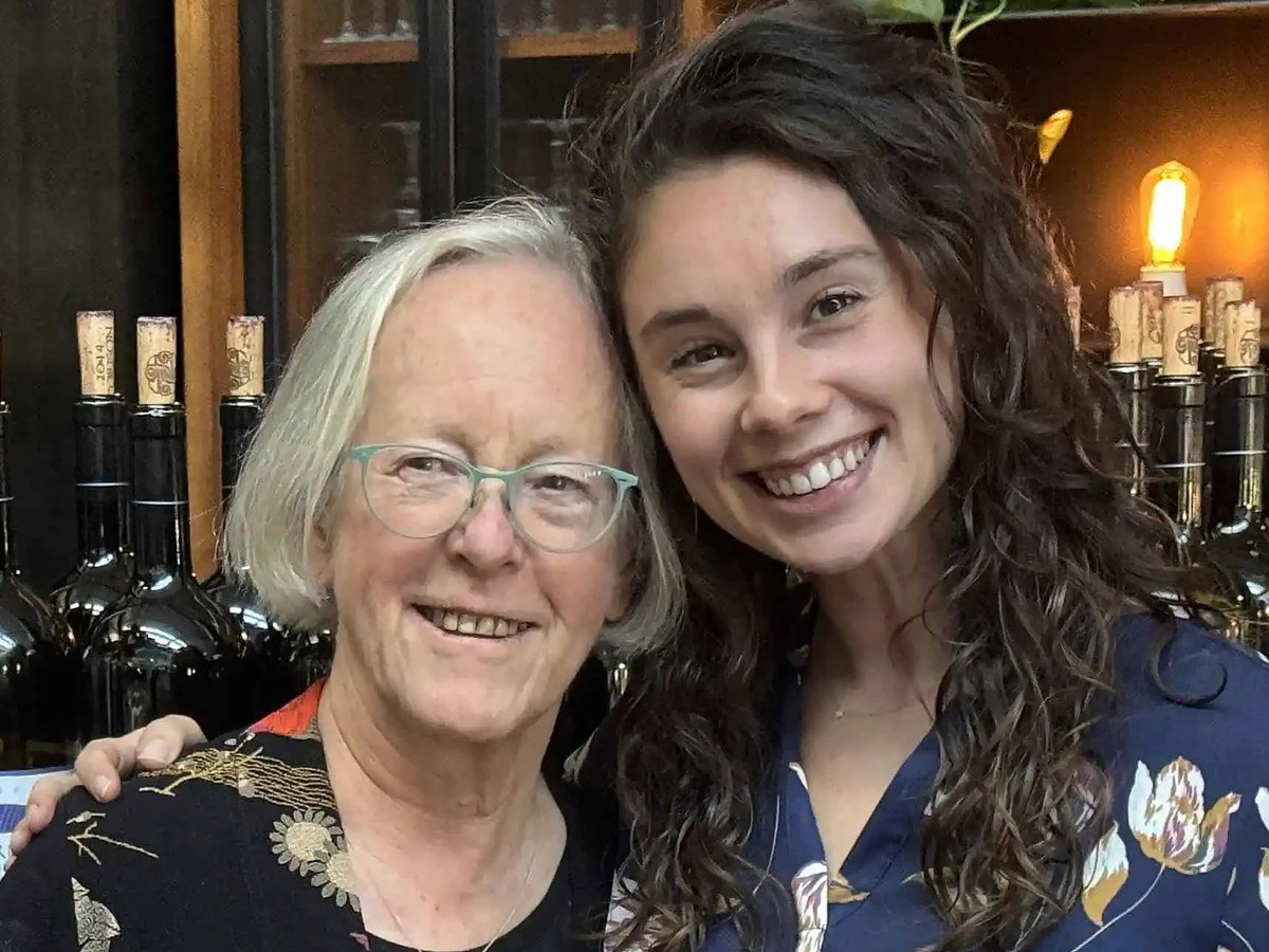 An older woman wearing glasses and a younger woman with long dark hair pose together indoors at what looks like a winery, with wine bottles on display behind them.