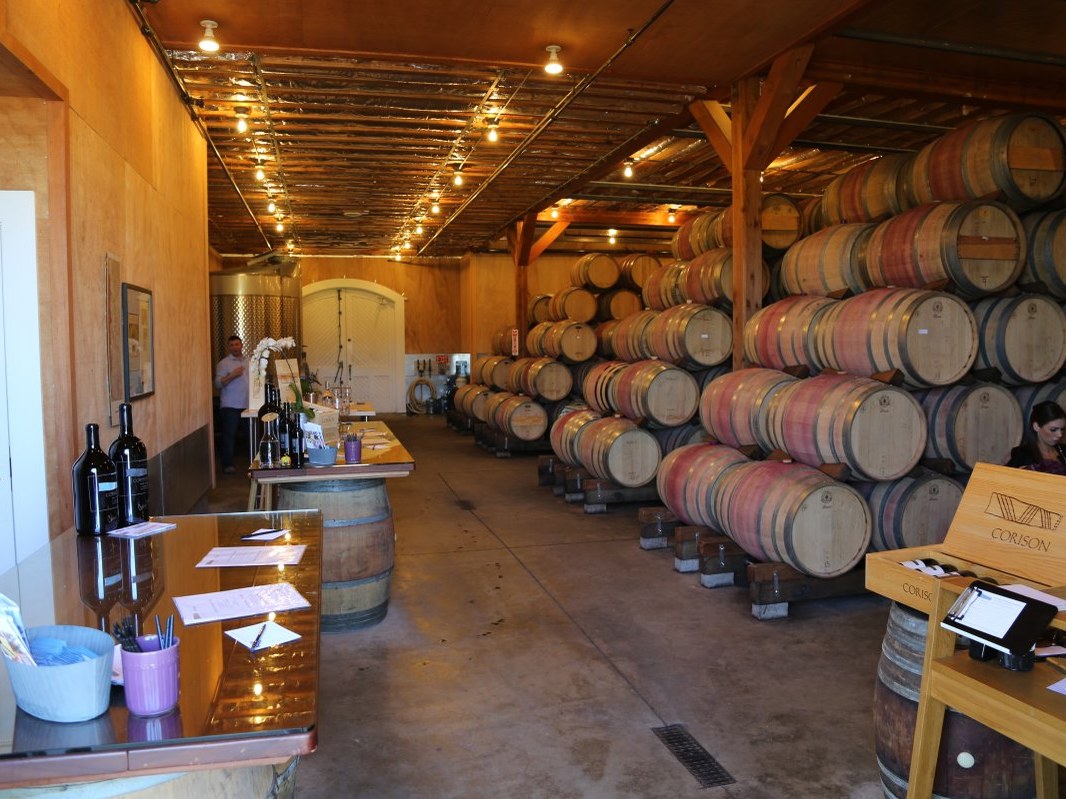 The interior of Corison Winery's barrel room, with rows of stacked oak barrels, a tasting counter, and wine bottles.
