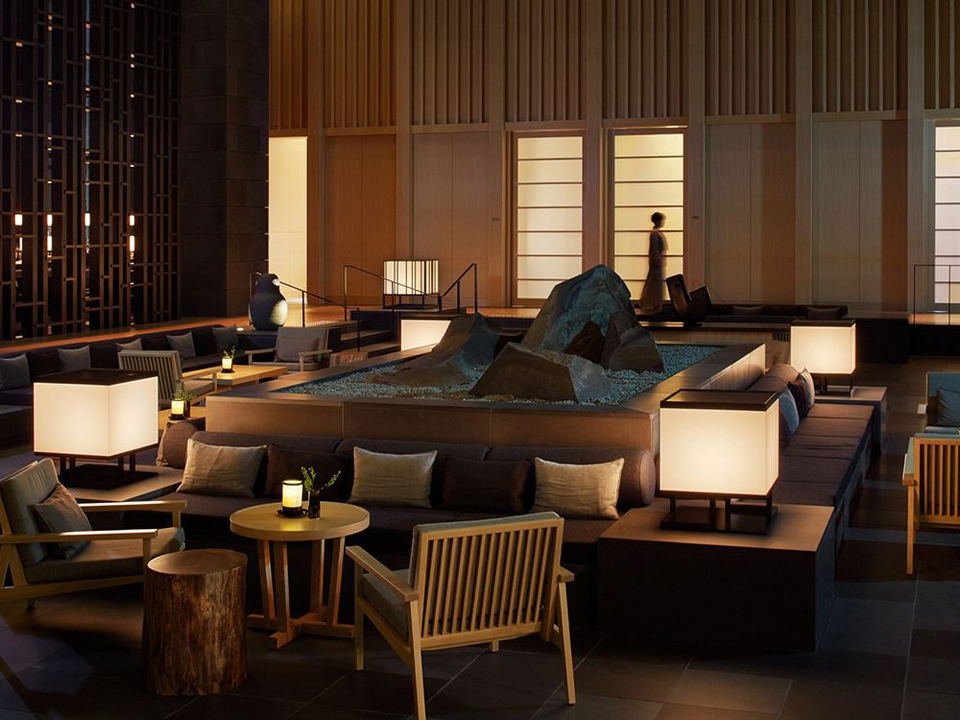 A warmly lit, Japanese-minimalist hotel lobby interior at Aman Tokyo, featuring a central rock garden, square glowing lanterns, low seating, and