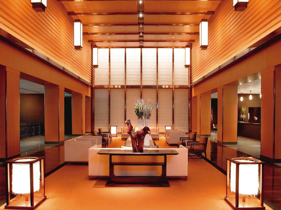 A warm, wood-paneled luxury hotel lobby with Japanese aesthetic design elements, lantern lighting, sculptural centerpiece, and staff visible at a