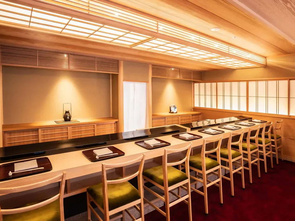 The interior of Sushi Yoshitake, featuring a long, light-colored hinoki wood counter with black lacquer accents, green-cushioned chairs, shoji screen