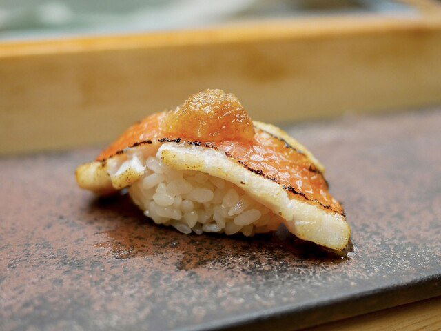 A close-up of a single piece of aburi nigiri sushi, featuring lightly torched fish on a bed of rice, served on a dark stone plate.