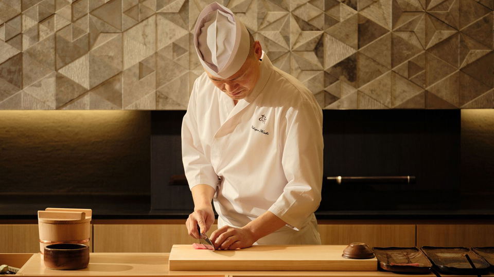 A sushi chef in traditional white uniform and tall chef's hat working at an intimate wooden counter, preparing sushi with precision; published by Cond