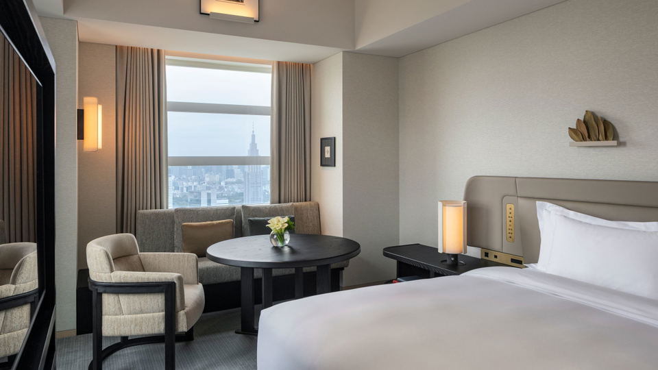 A modern luxury hotel guest room with floor-to-ceiling windows showing a city skyline view, featuring a bed with gray headboard, seating area with rou