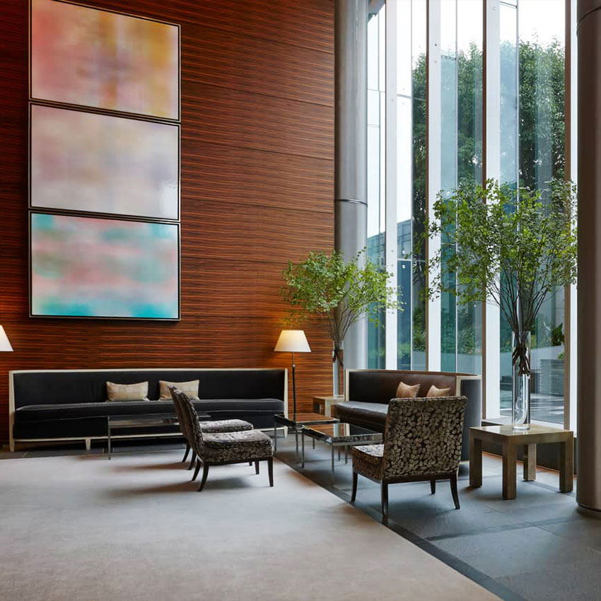 A refined hotel lobby with floor-to-ceiling windows, contemporary seating arrangements, abstract artwork on wood-paneled walls, and tall floral arrang