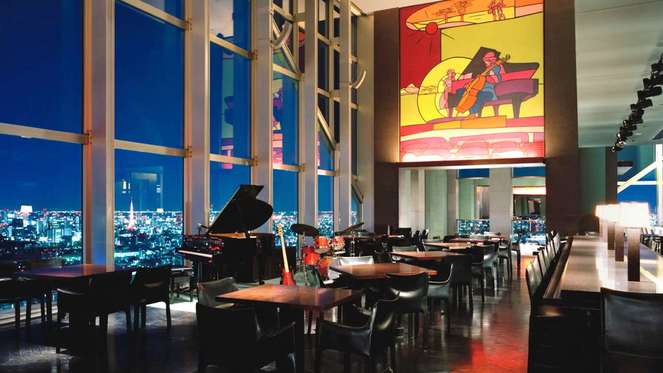 An upscale bar/lounge interior at night with floor-to-ceiling windows overlooking a sprawling city, featuring a grand piano, drum set, dining tables,