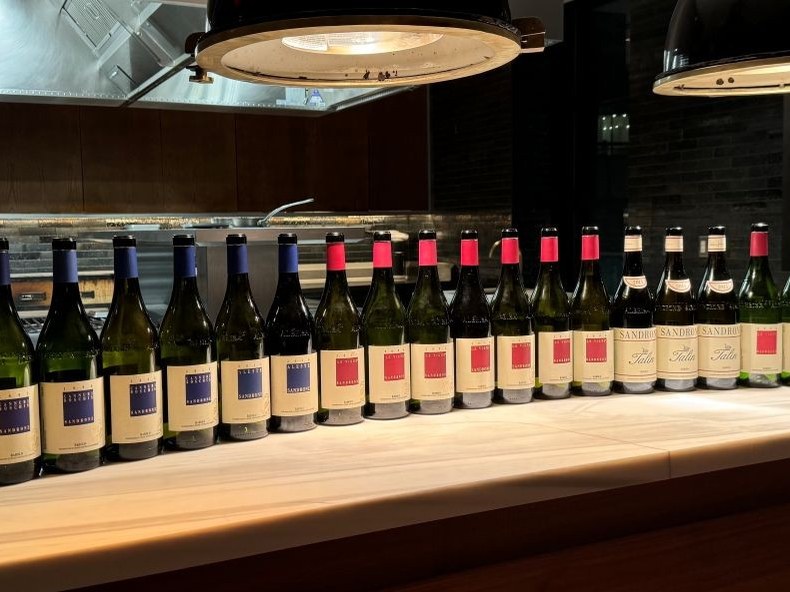 A long row of wine bottles with labels 'Sandrone', 'Cannubi Boschis', and 'Le Vigne' on a counter in a modern winery cellar.
