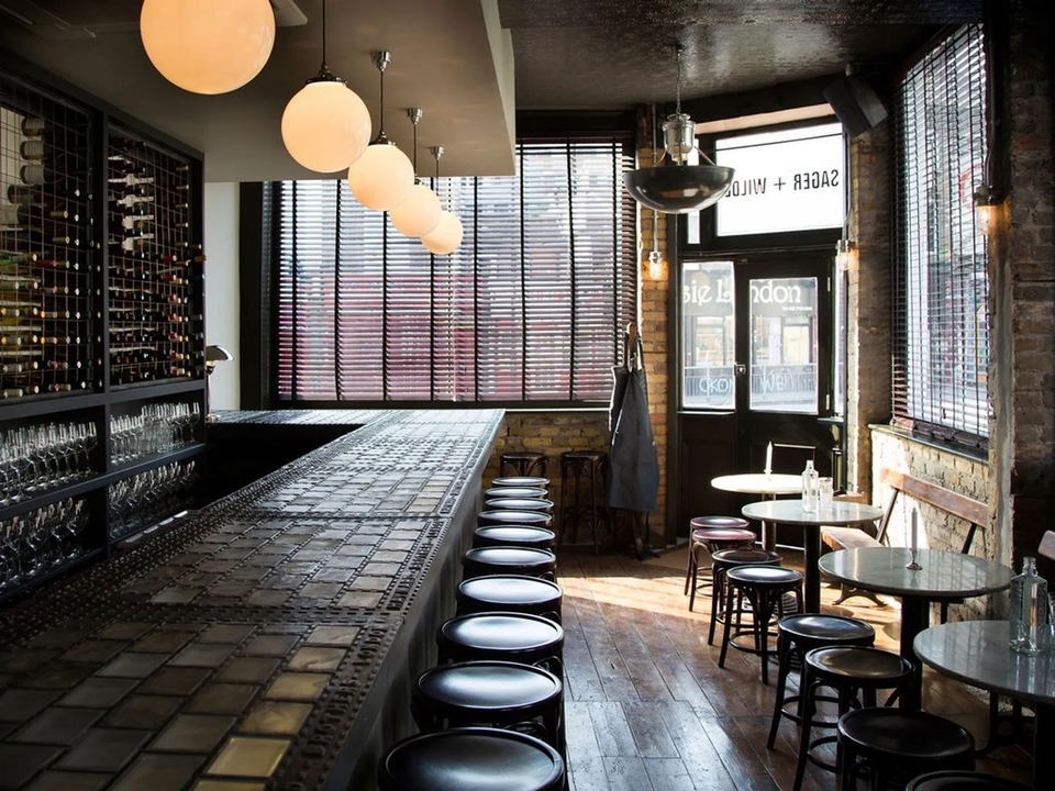 Interior of Sager + Wilde wine bar, featuring a long bar with a tiled top, rows of black bar stools, and globe pendant lights.