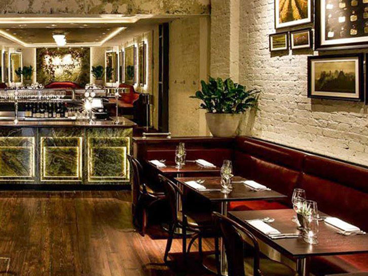 An elegant restaurant interior with dark wood floors, red leather banquette seating, a marble bar with green onyx panels, and framed vineyard artwork.