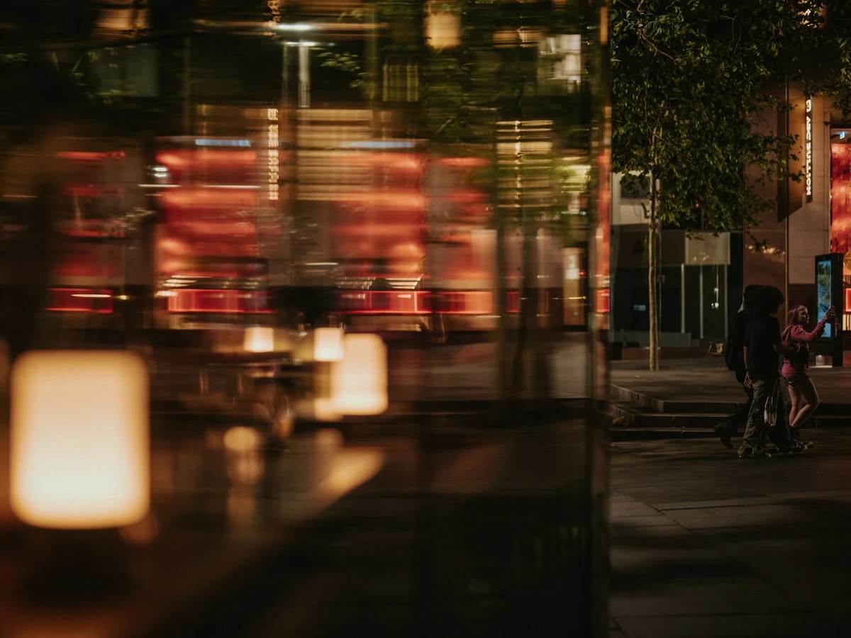 A motion-blurred red tram passes a building with floor-to-ceiling windows, revealing warm interior lighting. Two people walk on the pavement.
