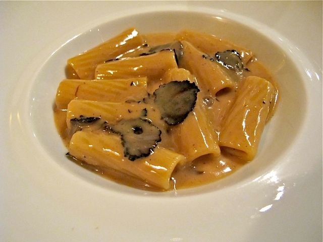 Close-up of ridged pasta tubes in a creamy sauce with black truffle shavings, plated in a white bowl; the source page caption explicitly identifies th