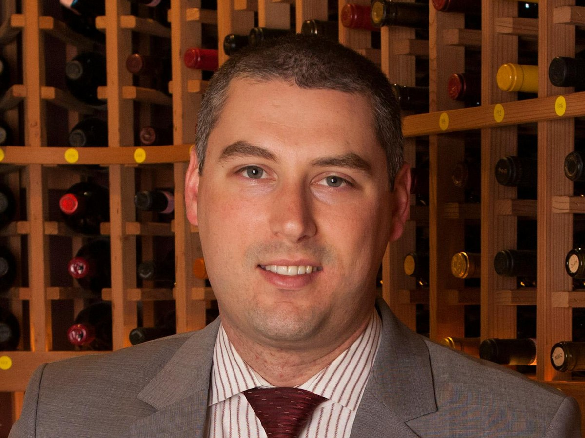 A man in a gray suit and striped shirt smiles at the camera while standing in front of a wooden wine rack filled with bottles.