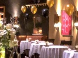 An upscale restaurant interior with white tablecloths, gold and black balloons, and warm lighting.