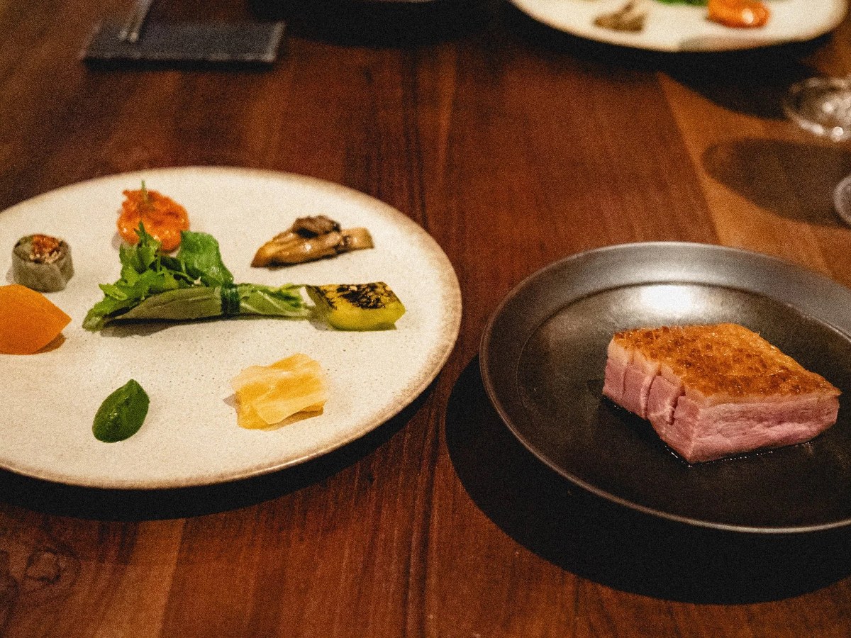 Two plated dishes from a fine-dining tasting menu: a white plate with multiple small composed elements (vegetables, proteins, garnishes) and a black p