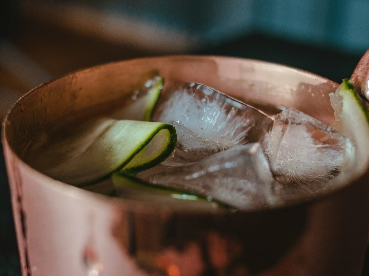 A pink-hued cocktail in a glass with ice cubes and thin cucumber slices as garnish, photographed against a blurred background.