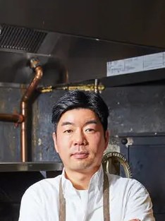 An Asian chef in a white chef's coat and apron stands in a professional kitchen with copper pipes and dark walls visible behind him.