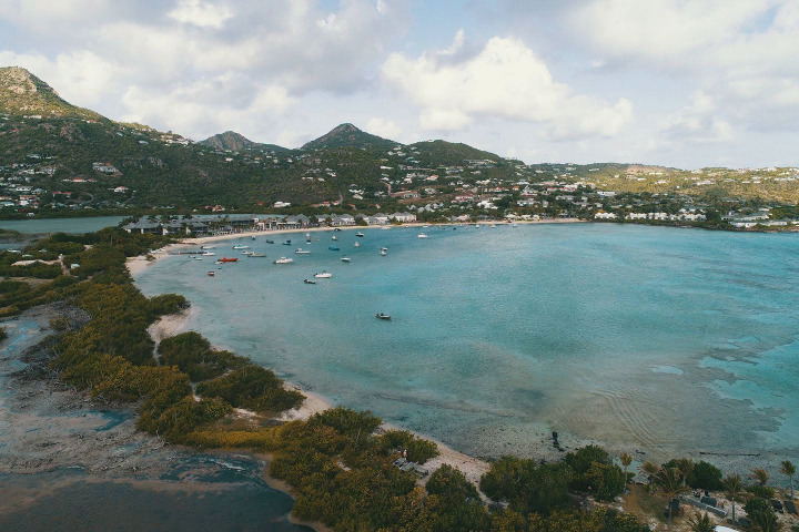Aerial view of a turquoise lagoon bay with boats, a peninsula with vegetation, and hillside development in Saint-Barth