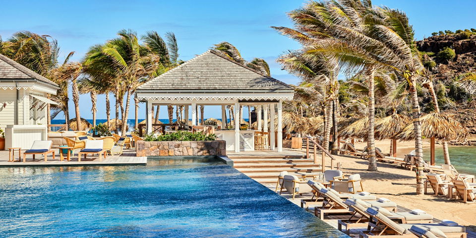 An infinity pool with white lounge chairs and a white pavilion structure overlooking a lagoon, surrounded by palm trees