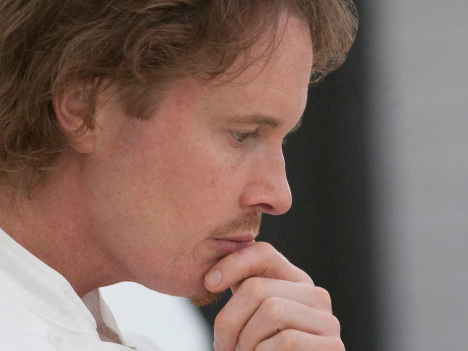 A man with brown hair and a light mustache, looks down thoughtfully, resting his chin on his hand.