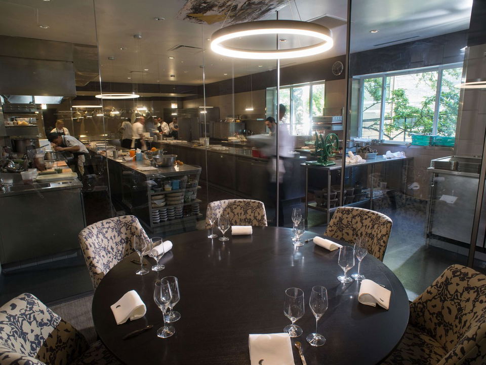 A round, dark wooden table is set with wine glasses and white napkins, positioned in front of an open kitchen where chefs are working.
