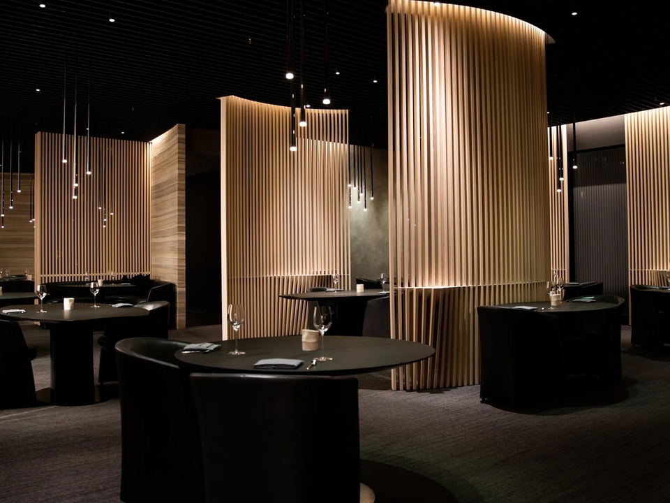A dimly lit fine-dining interior with vertical wood slat partitions, dark round tables, and pendant lighting.