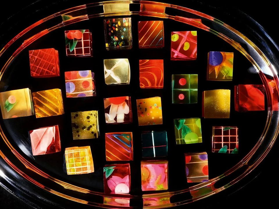 An overhead view of approximately 30 colorful, artistic bonbons or chocolates arranged on a black oval platter with a multicolored rim.