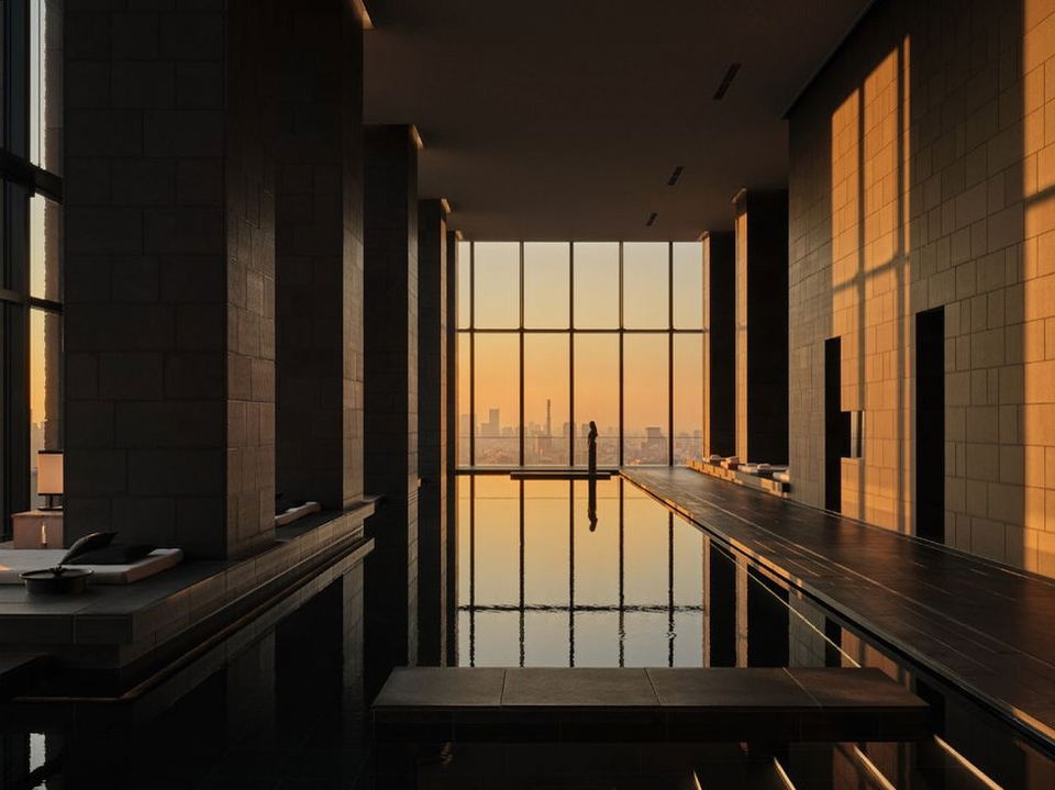 An indoor infinity pool at Aman Tokyo, with dark stone walls, floor-to-ceiling windows framing a golden-hour Tokyo cityscape, and a lone.