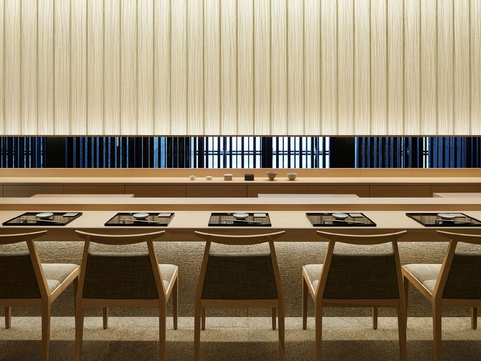 A minimalist Japanese sushi counter dining room with a light wood counter, black lacquer place settings, and a backlit washi-panel wall.