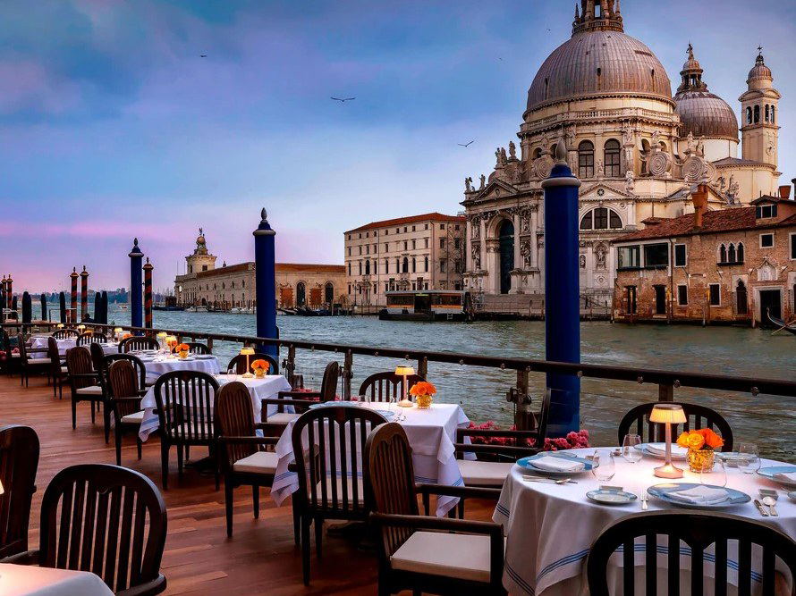 An elegant outdoor dining terrace at dusk, with tables set with white linens and lamps, overlooking the Grand Canal and the Basilica di Santa Maria.