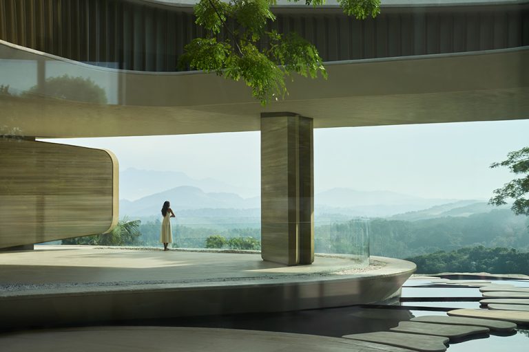 Architectural rendering of a contemporary circular platform with concrete flooring, wooden elements, and a person standing near floor-to-ceiling windows overlooking forested mountains.
