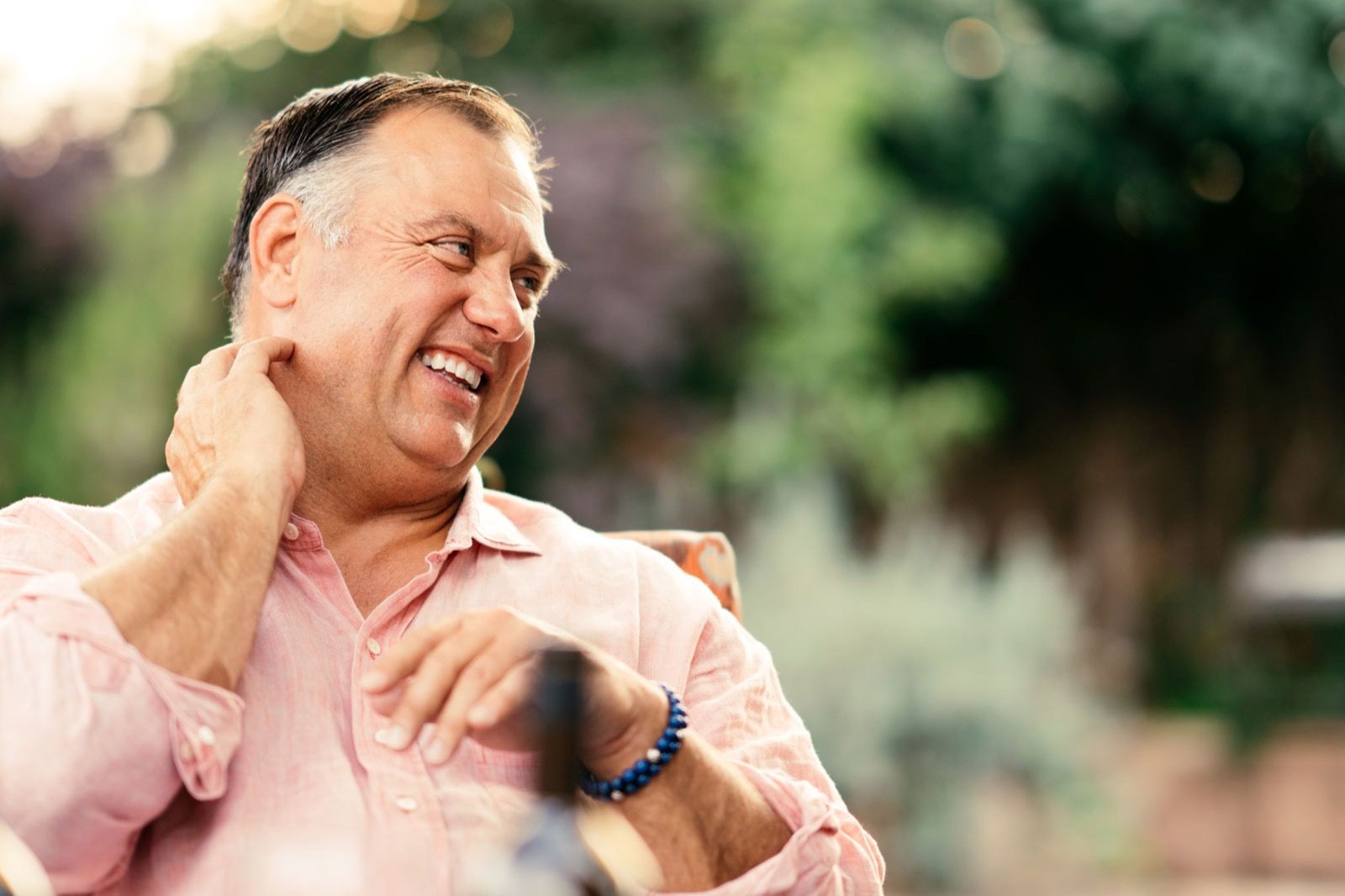 Tony Biagi, a man with short dark hair, smiles and scratches his neck while wearing a light pink shirt and a blue beaded bracelet.