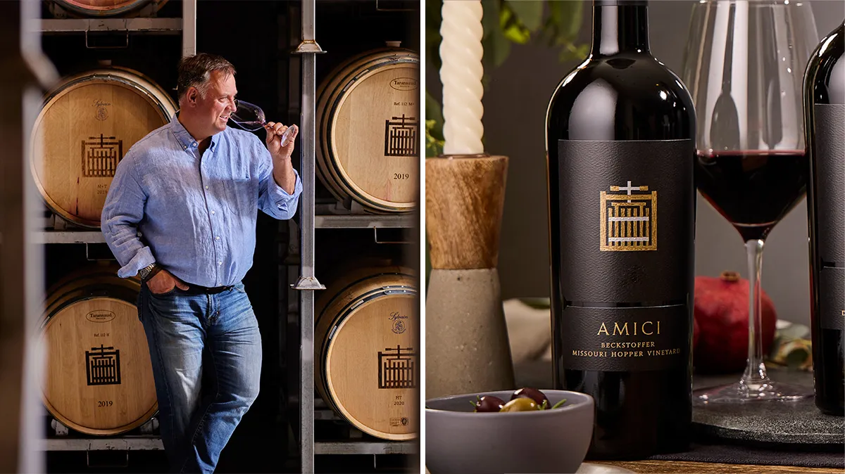 A man in a blue shirt and jeans holds a wine glass, smelling the wine, in a cellar with wooden barrels.