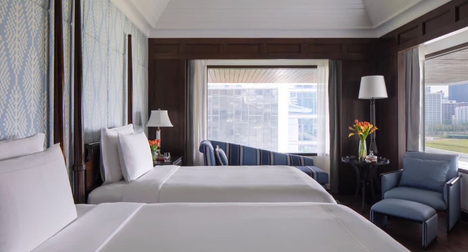 A luxury hotel bedroom with two beds featuring white linens, dark wood paneling, blue upholstered furniture, and Thai silk design elements.