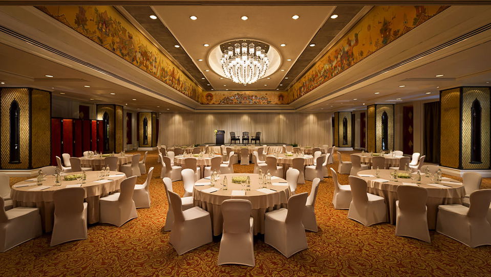 A luxurious hotel ballroom set for a banquet event with round tables, white chair covers, ornate ceiling with chandelier, and decorative murals; the s