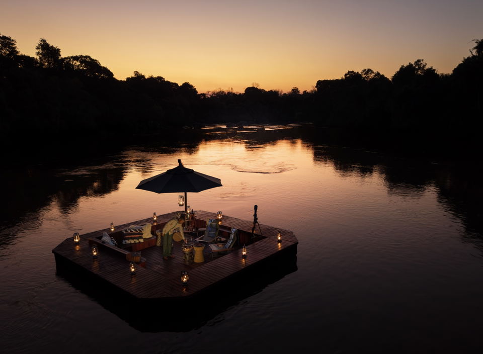 A floating wooden pontoon dining platform on a river at sunset, set with seating for four, lanterns, umbrella, and dining setup