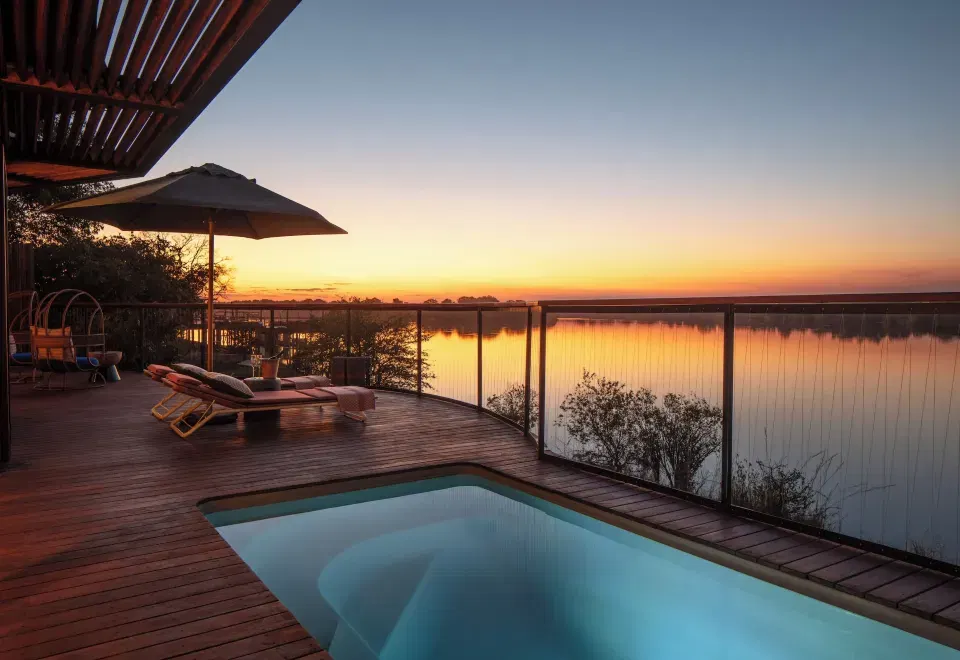 A luxury villa deck at sunset featuring a heated plunge pool on timber decking with glass railings overlooking a river, matching the Pool and Bush Vil