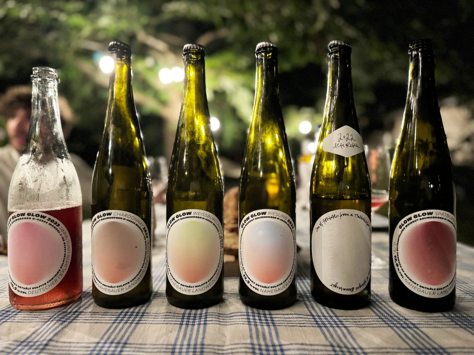 Six bottles arranged in a row on a striped tablecloth outdoors, showing various wine styles with circular labels identifying different varietals and styles from the Glow winery.