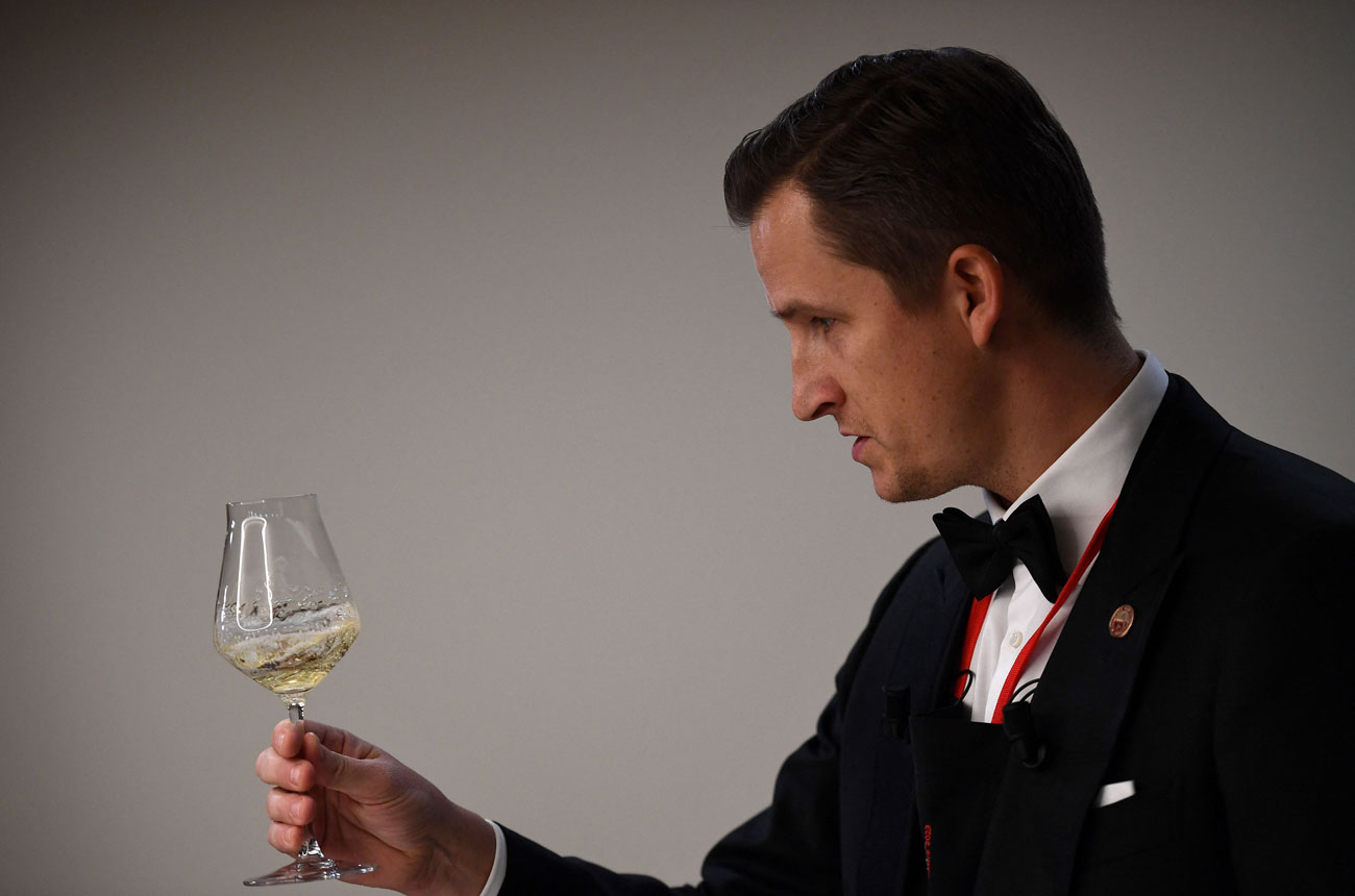 A man in formal attire (black tuxedo with bow tie and medal/ribbon) examining a glass of white wine in a professional tasting context