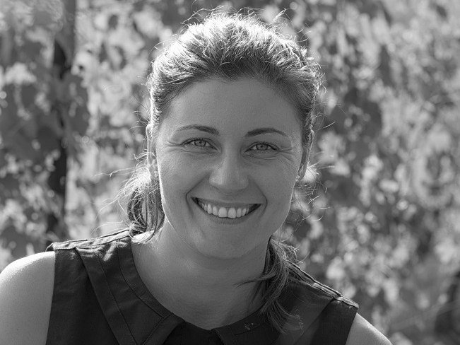 A smiling woman, Gaia Gaja, stands in a sunlit vineyard with blurred leaves in the foreground and background.