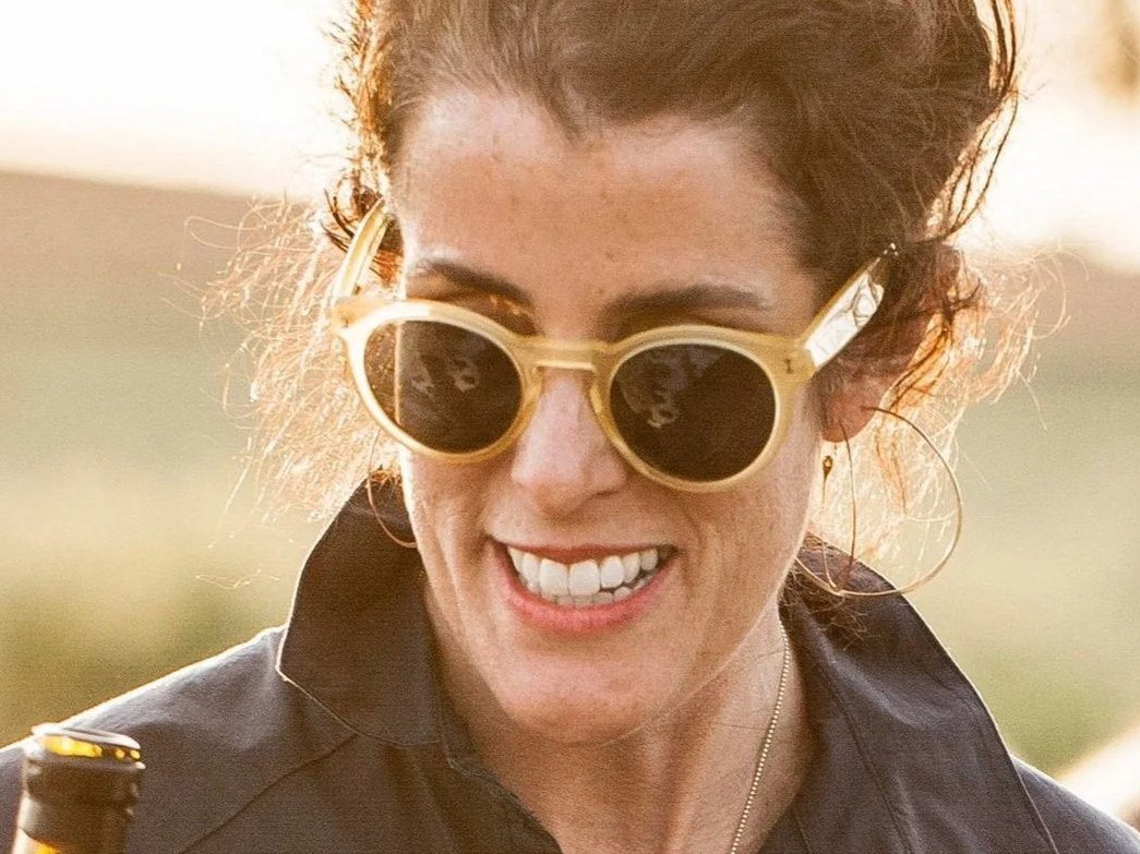 Maggie Harrison, with curly dark hair in an updo, smiles while wearing gold-framed sunglasses and a dark shirt, holding a wine bottle outdoors.