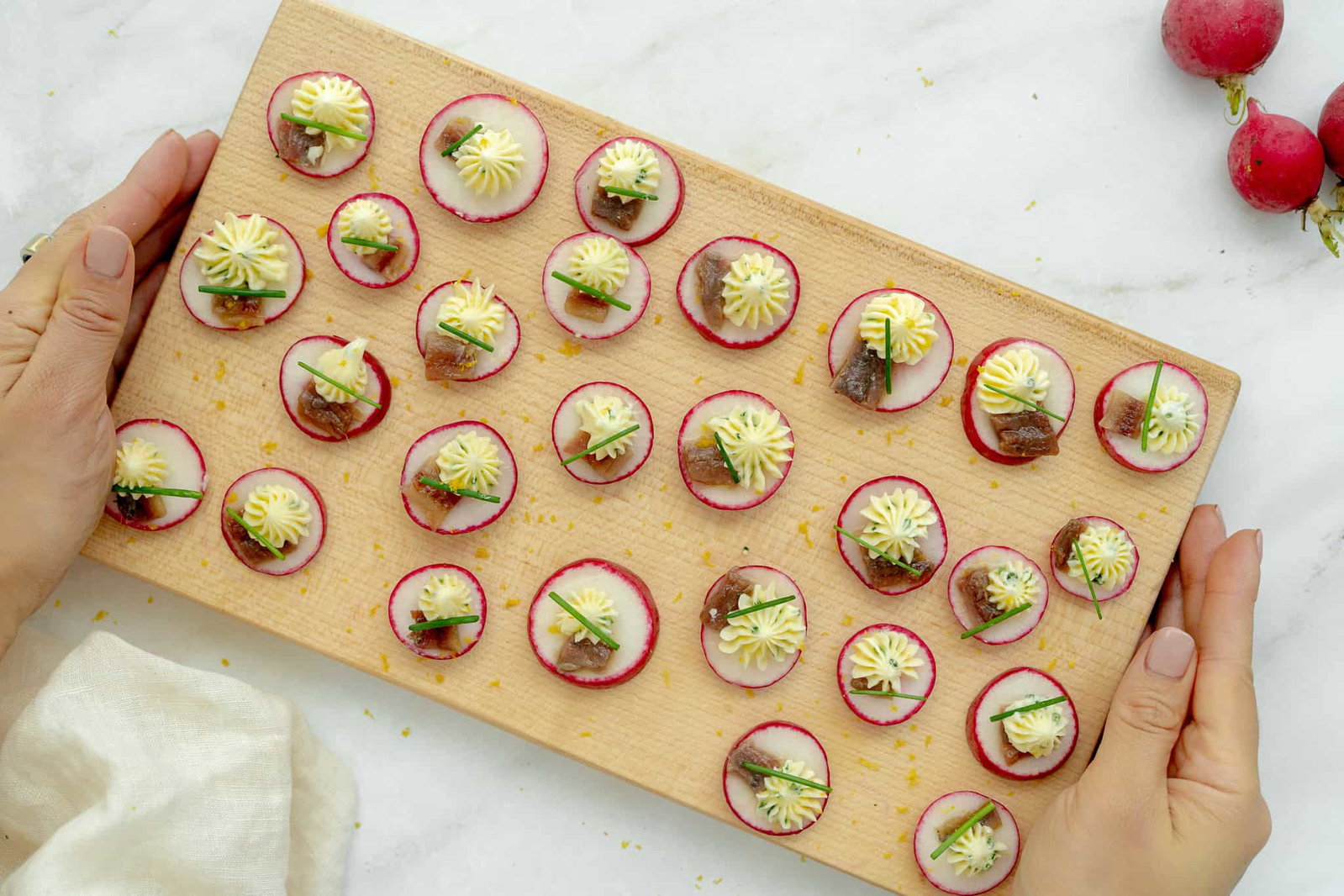 Radish slices topped with butter rosettes, anchovies, and chive garnish are arranged on a wooden serving board held by two hands.