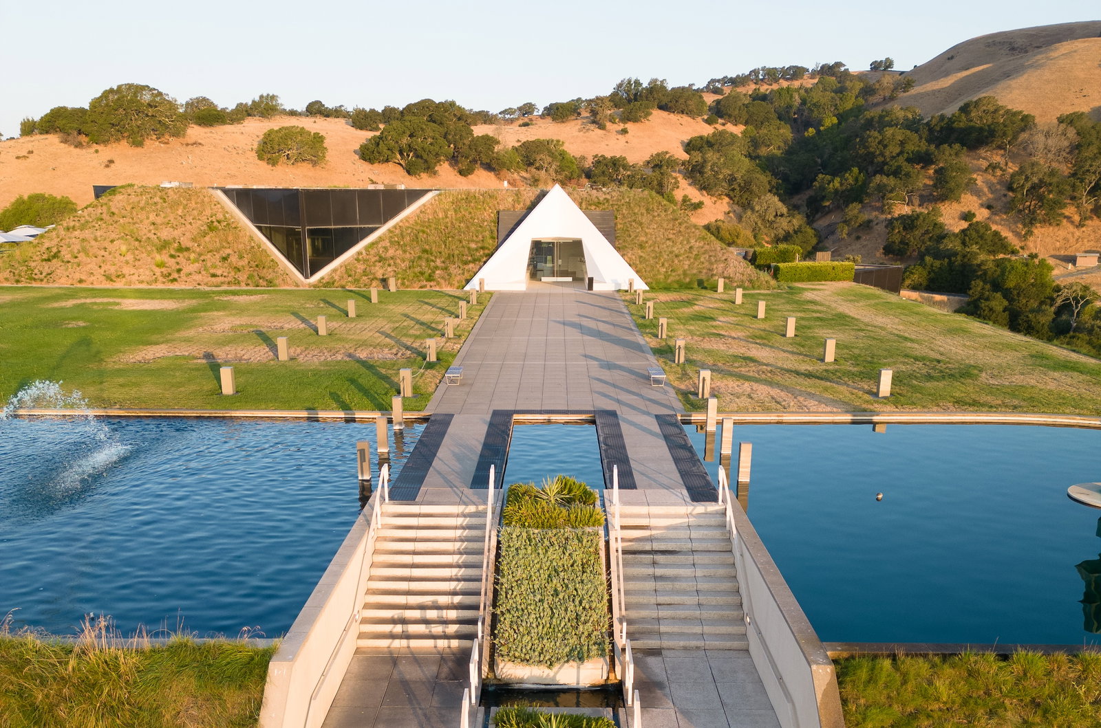 Aerial view of a modern winery with distinctive pyramid-shaped architecture set into hillside vineyards, with a reflecting pool and formal walkway; so