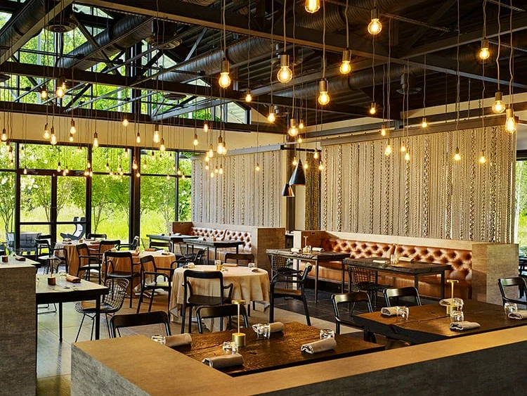 A spacious, modern restaurant with high ceilings, exposed ductwork, and numerous hanging Edison bulbs, featuring a mix of dining tables and booths.